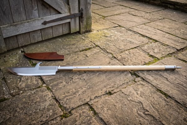 Hand forged glaive polearm with a broad hooked single edged head and knotwork engraved socket on a long ash haft, lying on flagstones beside a brown leather sheath.