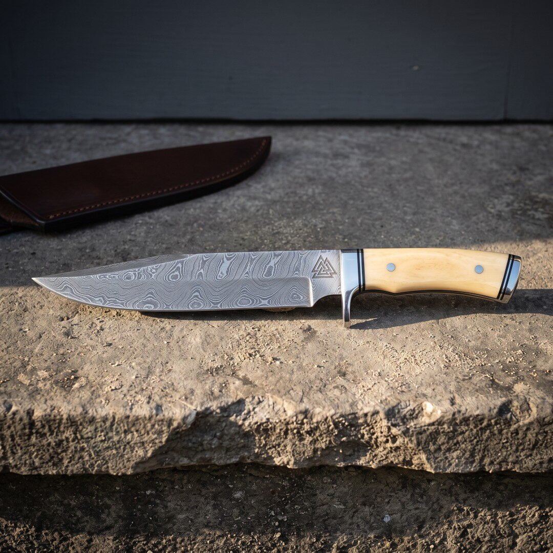 Gemini_Generated_Image_uvoo6nuvoo6nuvoo Hand forged damascus viking bowie knife with pale bone handle scales and a Valknut etched blade resting on rough stone beside its oxblood leather belt sheath.