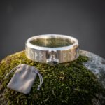 Hand made viking rune ring in polished steel with raised Elder Futhark characters and an applied Mjolnir hammer, standing on green moss beside a grey suede pouch