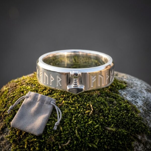 Hand made viking rune ring in polished steel with raised Elder Futhark characters and an applied Mjolnir hammer, standing on green moss beside a grey suede pouch