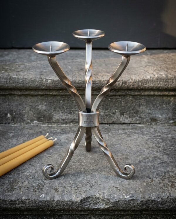 Hand forged viking candle holder in twisted brushed steel with three pricket branches, Mjolnir engraved collar and scrolled feet, on a stone step beside beeswax tapers