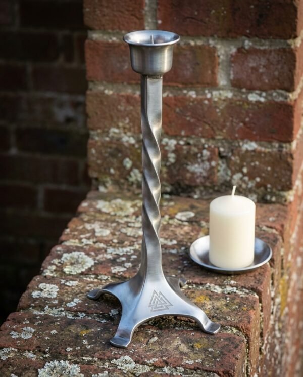 Hand made steel viking candle holder with a hand twisted stem and a Valknut engraved tripod foot, standing on a lichened brick wall beside a white pillar candle.