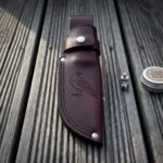 Hand made oxblood leather knife sheath with a tooled raven panel and steel snap retention strap, lying on weathered grey decking beside spare studs and a tin of leather balm.