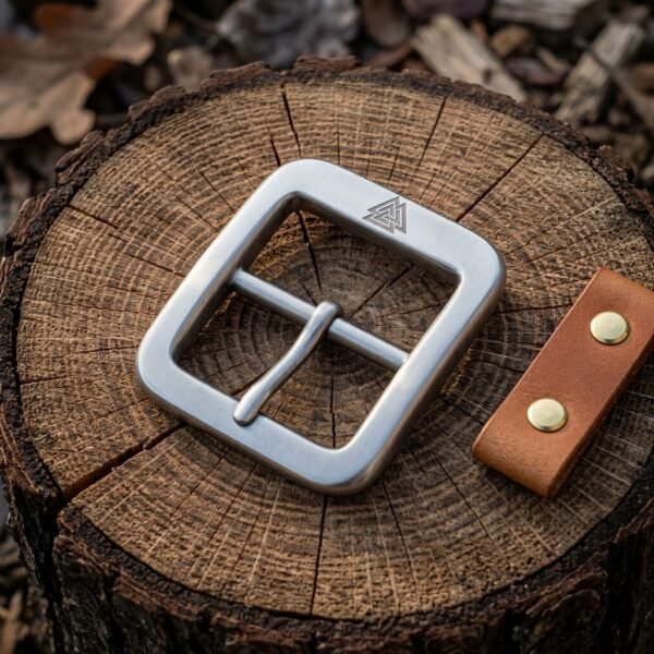 Hand made viking belt buckle in brushed steel with a stamped Valknut and centre bar tongue, on a cut tree stump beside a tan leather snap tab