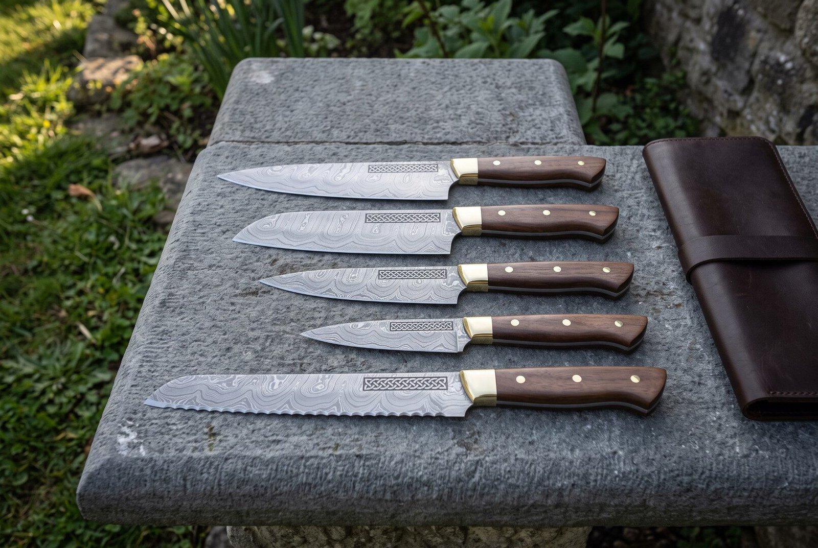 Gemini_Generated_Image_whh44awhh44awhh4 Hand forged Viking chef knife set of five Damascus blades with knotwork engraved panels, walnut handles and brass bolsters, laid on slate beside a brown leather knife roll.