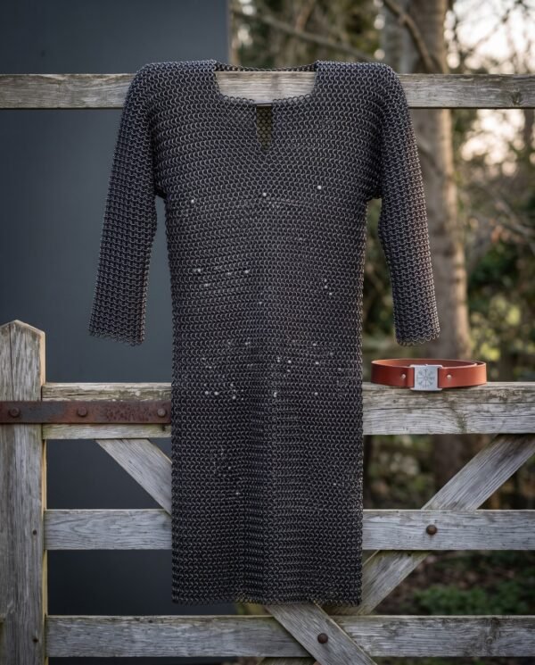Hand woven chainmail hauberk in blackened mild steel with a four in one butted weave and keyhole neck, hanging on a weathered timber gate in winter light