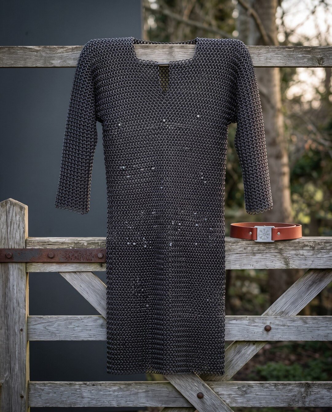 Gemini_Generated_Image_wkmu39wkmu39wkmu Hand woven chainmail hauberk in blackened mild steel with a four in one butted weave and keyhole neck, hanging on a weathered timber gate in winter light