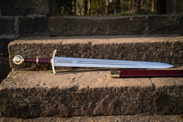 Hand forged medieval sword with a brass wheel pommel, oxblood leather grip and a knotwork etched blade, lying on a sunlit stone step beside its brass mounted scabbard.
