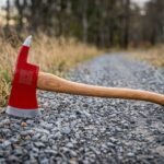 Hand forged felling axe with a red enamelled head, engraved Celtic knotwork, polished cutting bevel and hickory handle, standing on a gravel forest track in autumn.