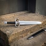 Hand forged fantasy sword with a twisted loop guard, black leather grip and polished steel wheel pommel, lying on a stone step beside its black scabbard with a steel chape.