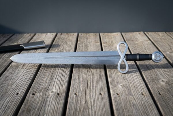 Hand forged damascus sword with an infinity loop guard, Mjolnir engraved pommel and black leather grip, laid on weathered grey decking beside a steel scabbard throat.