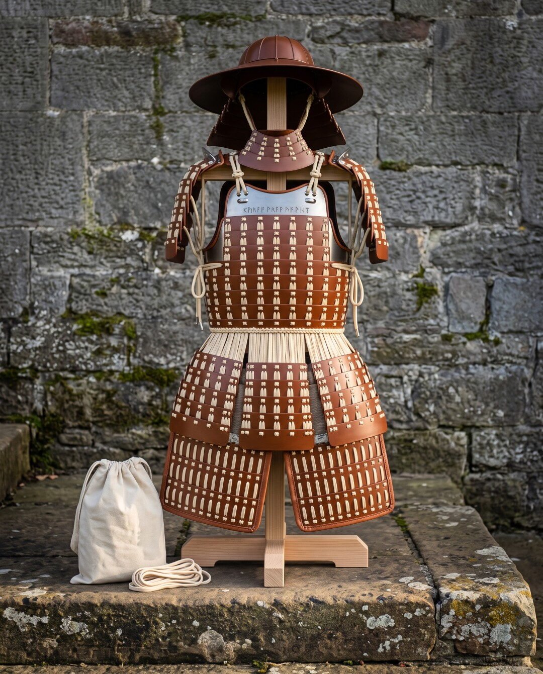 Gemini_Generated_Image_z9ouv9z9ouv9z9ou Hand laced leather lamellar samurai armor on a wooden stand against a stone wall, showing wide brim helmet, shoulder guards, rune engraved chest plate and thigh guards.