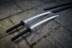 Angled overhead view of the twin ninjato set on grey flagstones, showing both polished blades, the black cord wrapped handles and the pair of matching black scabbards.