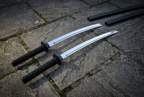Angled overhead view of the twin ninjato set on grey flagstones, showing both polished blades, the black cord wrapped handles and the pair of matching black scabbards.