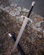 Angled view of a fluted pommel damascus longsword resting on a lichen covered wall, showing the faceted steel crown, black leather grip and knotwork panel on the guard