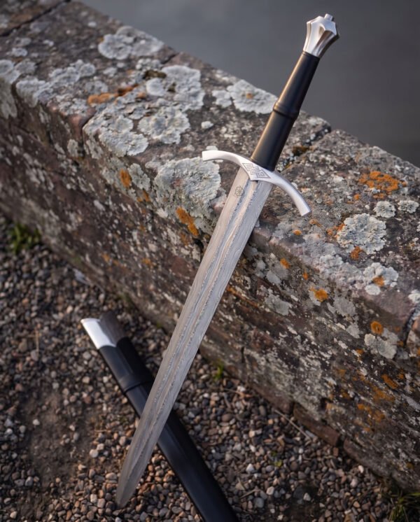 Angled view of a fluted pommel damascus longsword resting on a lichen covered wall, showing the faceted steel crown, black leather grip and knotwork panel on the guard