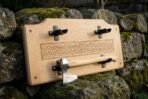 Angled view of a forged axe resting in black cradles on a carved oak knotwork plaque against a mossy dry stone wall