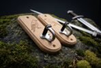 Angled view of matched oak sword hanger plaques with raven engravings and steel hooks, two swords lying behind