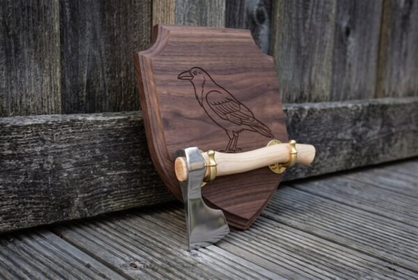 Angled view of a raven engraved walnut wall plaque with a polished axe and pale ash haft held in brass cradle brackets