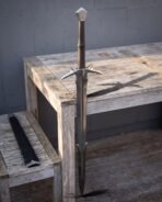 Angled view of a serrated fantasy sword and its black scabbard on rough sawn wood, barbed crossguard and notched blade shoulders catching the afternoon light