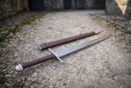 Angled view of the knotwork panel damascus sword and brown leather scabbard on a stone courtyard floor, showing the layered blade pattern and the etched panel above the ricasso.