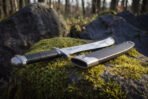 Angled view of the polished sawback short sword resting on green moss beside its black sheath, with the steel throat, chape and bright upswept blade in sunlight.