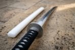 Antiqued steel tsuba with Norse knotwork banding shown close up over the black cord wrapped tsuka of a wakizashi beside its gloss white lacquered scabbard