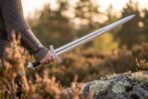 Arm in mail and tooled leather bracer holding a polished arming sword with serpent etched fuller over heather at sunset