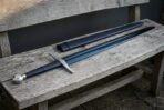 Arming sword with blued blade, wheel pommel and black leather grip laid full length on a grey wooden bench next to its scabbard