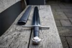 Close up of an arming sword hilt with wheel pommel, black leather grip and steel cross guard on a weathered bench, blued blade beyond