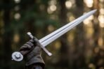 Arming sword with an engraved crossguard and wheel pommel held up in a gloved hand against a soft forest background
