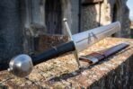 Arming sword with black leather grip and round wheel pommel resting on a brick wall beside its brown leather scabbard