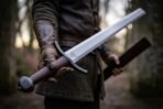 Arming sword with blackened curved guard, oxblood leather grip and faceted pommel held in a gloved hand