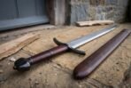 Arming sword with oxblood leather grip and blackened faceted pommel lying beside its matching scabbard on stone