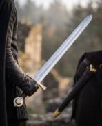 Arming sword with rune cut fuller and brass wheel pommel held in a gloved hand near ruins, black scabbard in the background