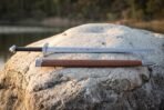 Arming sword with serpent etched fuller and black grip lying on a boulder by a lake above its brown leather scabbard