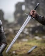 Armoured hand raising a Damascus steel hand and a half sword with interlace panel and black grip in a misty ruin