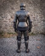 Back view of a blackened steel plate armour suit showing fluted backplate, rune line and quilted skirt by a stone wall