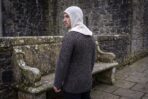 Back view of a man wearing a blackened steel chainmail shirt and a cream linen coif beside a stone bench in a castle yard
