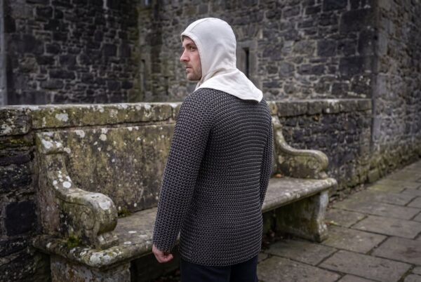 Back view of a man wearing a blackened steel chainmail shirt and a cream linen coif beside a stone bench in a castle yard