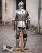 Back view of a steel plate armour harness on a wooden stand showing the backplate, leather straps and leg harness