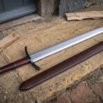 Ball pommel arming sword in satin 1095 steel with oxblood leather grip and blackened curved guard lying on a stone doorstep beside its matching leather scabbard