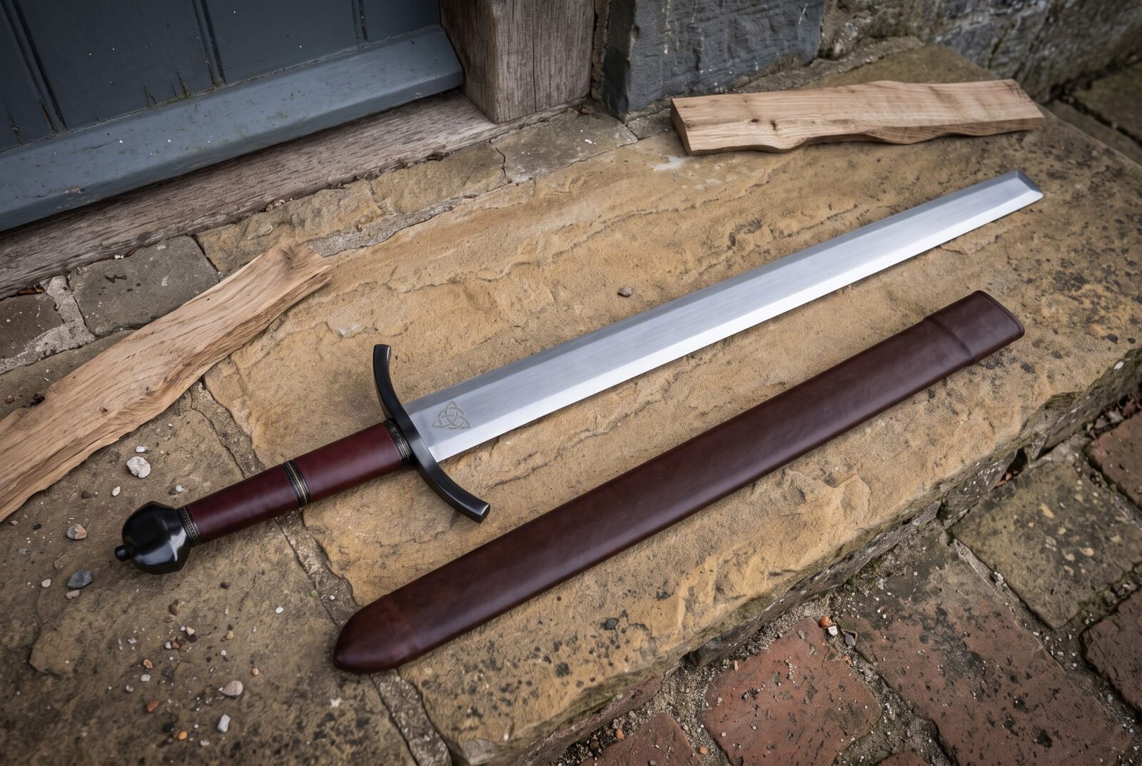Ball pommel arming sword in satin 1095 steel with oxblood leather grip and blackened curved guard lying on a stone doors Ball pommel arming sword in satin 1095 steel with oxblood leather grip and blackened curved guard lying on a stone doorstep beside its matching leather scabbard