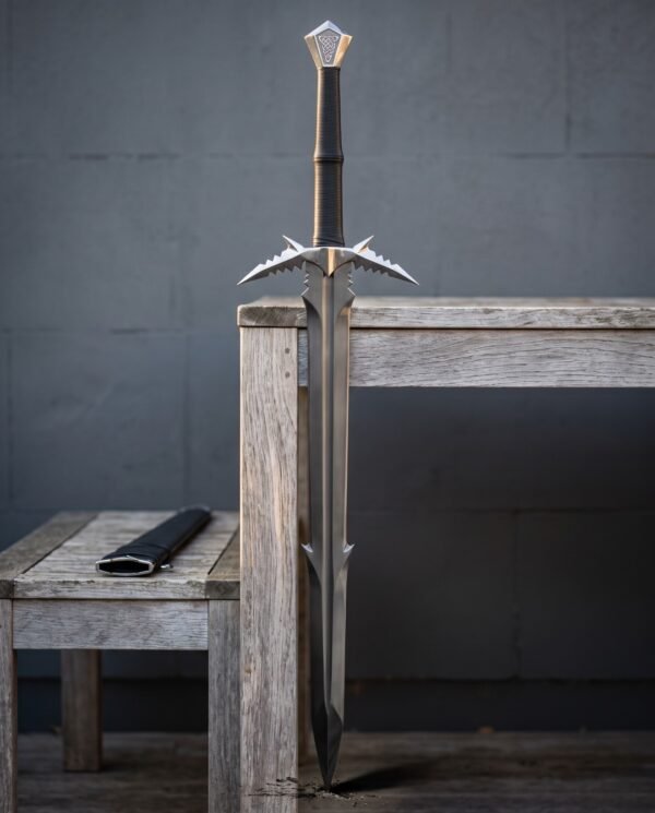 Barbed fantasy sword standing point down on a weathered wooden bench showing its spiked crossguard, knotwork pommel and black scabbard against a grey wall