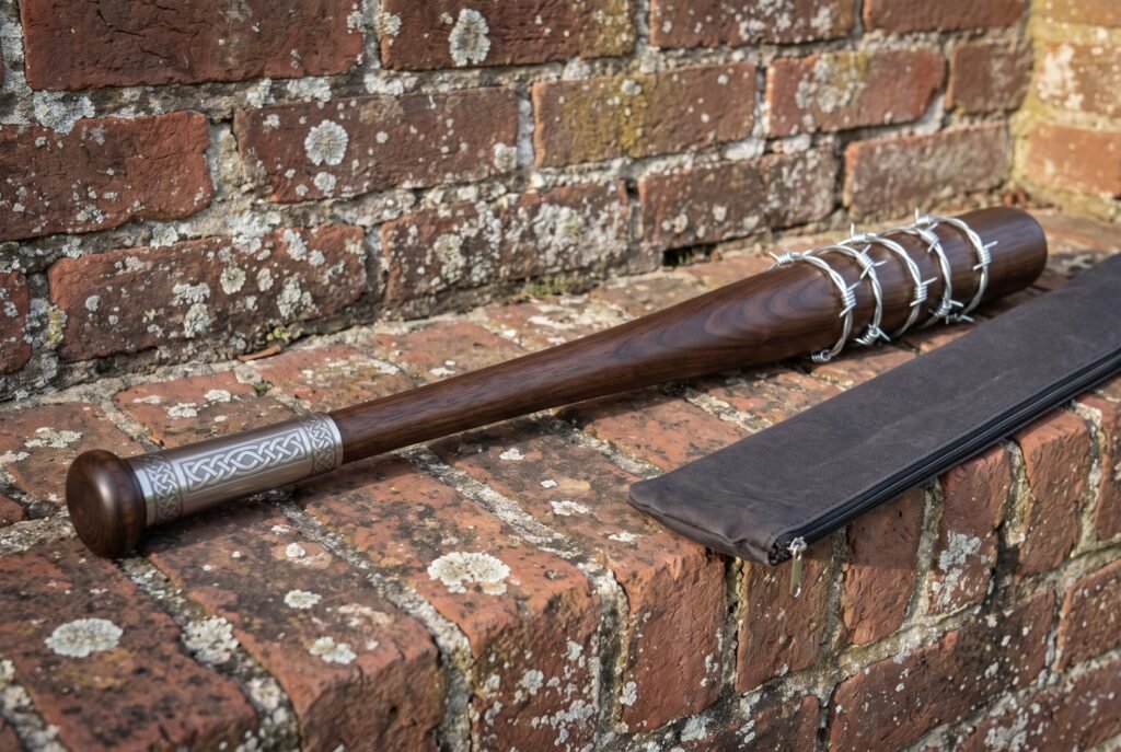 Barbed wire baseball bat in turned hardwood with an engraved steel collar and knob resting on a weathered red brick wall beside its carry case