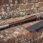 Barbed wire baseball bat in turned hardwood with an engraved steel collar and knob resting on a weathered red brick wall beside its carry case
