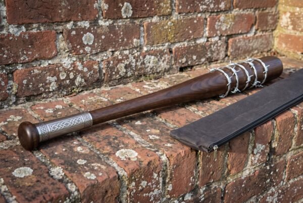 Barbed wire baseball bat in turned hardwood with an engraved steel collar and knob resting on a weathered red brick wall beside its carry case