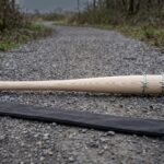 Barbed wire bat turned from pale ash with a galvanised wire wrap and engraved steel butt cap lying on a gravel track beside its black canvas carry sleeve
