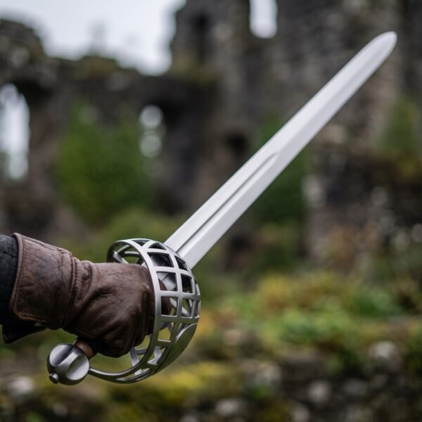 Basket hilt sword with steel bar cage held in a brown leather glove in front of blurred castle ruins