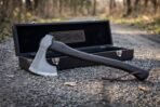 Bearded axe with dark hardwood haft and leather wrist thong resting against its open black presentation case on a gravel path